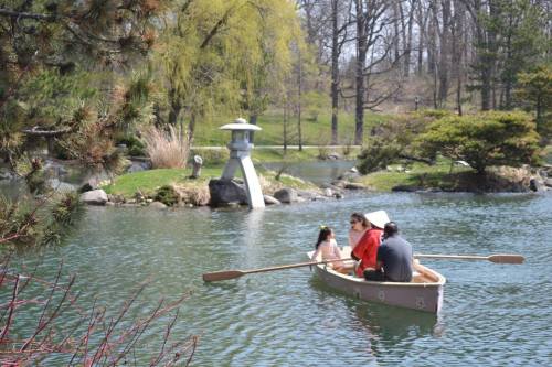 A family rows a small boat on a serene pond surrounded by greenery and a lantern in the background.