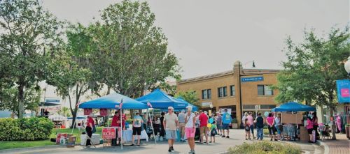 A lively outdoor market scene with people browsing stalls under blue tents, surrounded by trees and buildings.