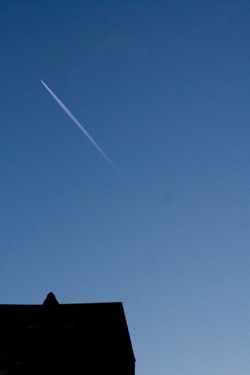 A clear blue sky with a single contrail streaking across, above the silhouette of a house.