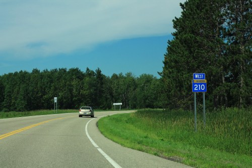 A car drives on a winding road with a sign indicating "West 210" and lush green trees in the background.