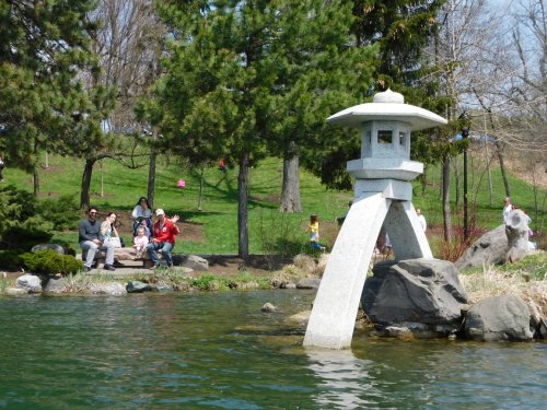 A stone lantern stands by a pond, with people sitting on a bench and enjoying the park's greenery in the background.