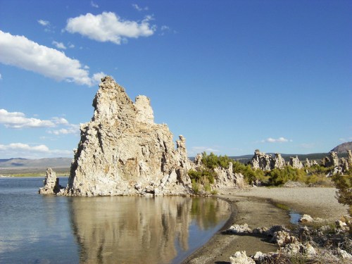 Rocky formations rise from the water's edge under a blue sky with scattered clouds, reflecting in the calm lake.