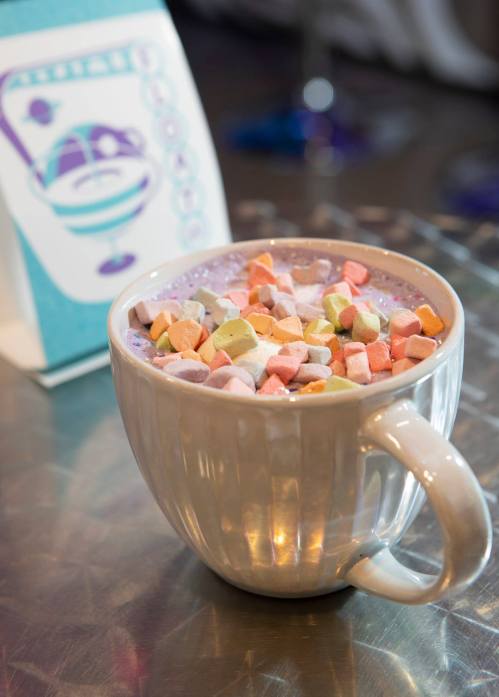 A mug filled with hot chocolate topped with colorful marshmallows, next to a retro-style menu card.