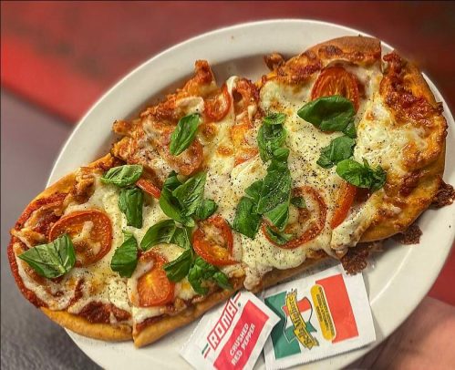 A plate of flatbread pizza topped with cheese, tomatoes, and fresh basil, served with red pepper flakes and seasoning packets.