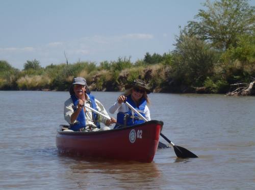 Two people paddle a red canoe on a calm river, surrounded by greenery and a clear blue sky.