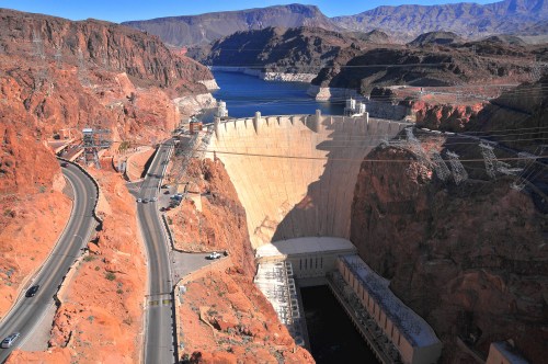 Aerial view of a large dam surrounded by rocky terrain and a river, with power lines in the background.