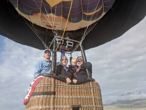 A family smiles from a hot air balloon basket against a cloudy sky.