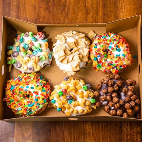 A box of six colorful donuts topped with various cereals and candies, arranged on a wooden surface.