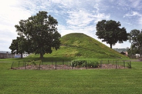 A grassy mound surrounded by trees, with a garden enclosed by a fence in the foreground under a cloudy sky.