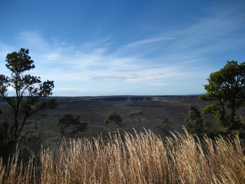 A panoramic view of a volcanic landscape with tall grass and sparse trees under a blue sky.