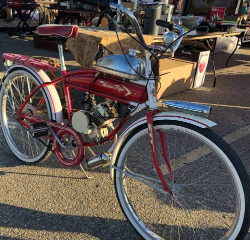 A vintage red and white motorized bicycle with a chrome engine, parked at an outdoor market.