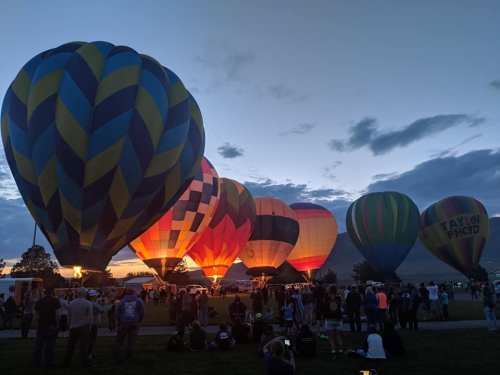 A colorful array of hot air balloons illuminated at dusk, with a crowd gathered below in a grassy field.