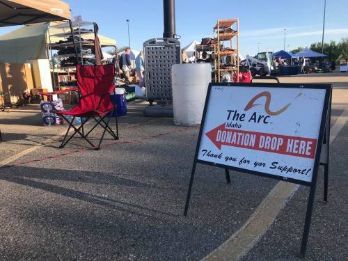 A sign for The Arc Idaho indicating a donation drop-off location, with a red folding chair nearby at an outdoor event.