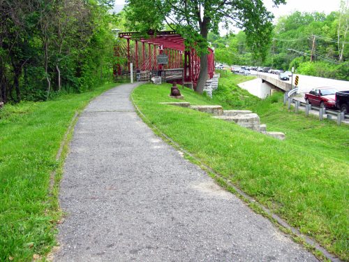 A paved path lined with grass leads to a red structure, with a road and cars visible in the background.
