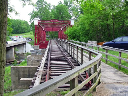 A red metal bridge spans a river, with a wooden walkway alongside old railway tracks and greenery in the background.