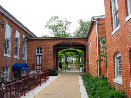 A brick walkway leads through an archway, flanked by buildings and greenery, with people in the distance.