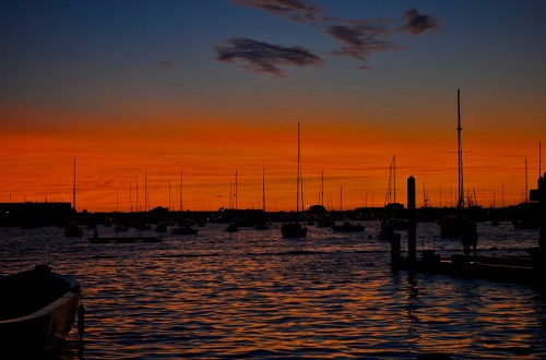 Silhouette of sailboats on water at sunset, with vibrant orange and blue sky reflecting on the surface.