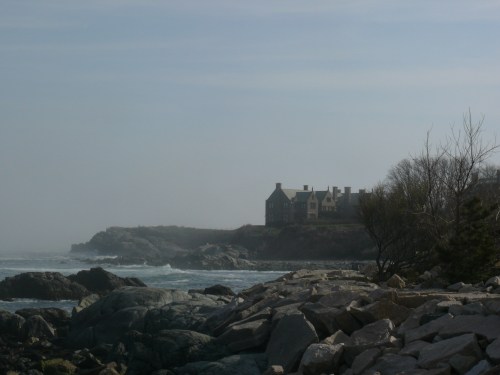A rocky shoreline with a foggy view of a large house on a cliff, surrounded by ocean waves and sparse vegetation.