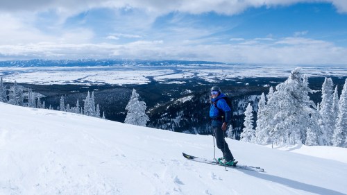 A skier stands on a snowy slope, surrounded by trees and mountains under a blue sky with scattered clouds.