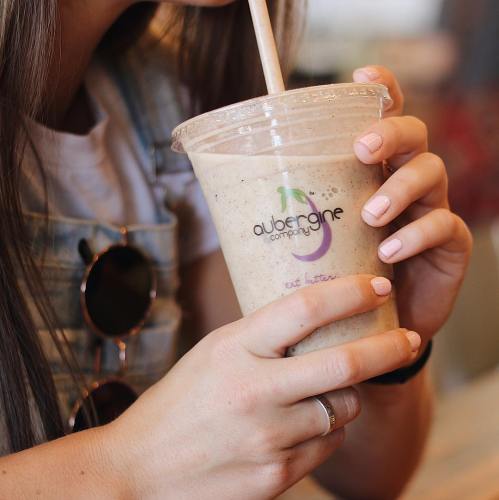 A close-up of a person holding a smoothie in a clear cup with a logo, wearing a denim outfit and sunglasses.