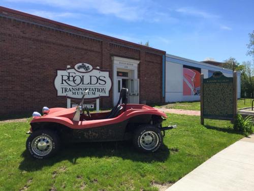 A red dune buggy parked outside the Reolds Transportation Museum, with a historical sign nearby and a brick building in the background.