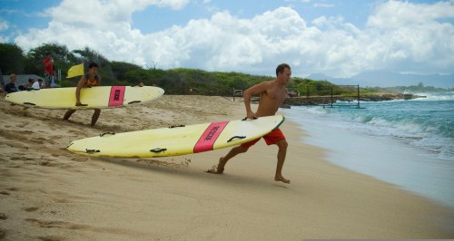 Two surfers run along the beach with yellow surfboards, preparing to catch waves under a partly cloudy sky.