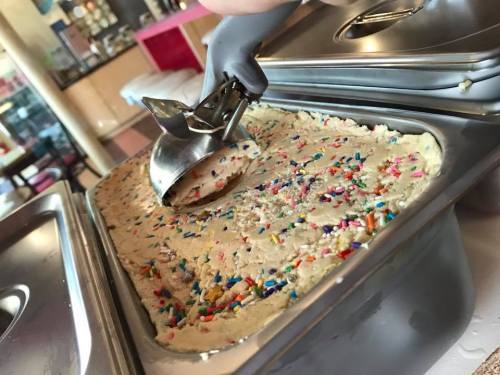 A hand using an ice cream scoop to serve colorful sprinkles-topped ice cream from a metal container.