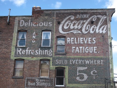 Faded Coca-Cola advertisement on a brick wall, featuring text about refreshment and pricing at 5 cents.
