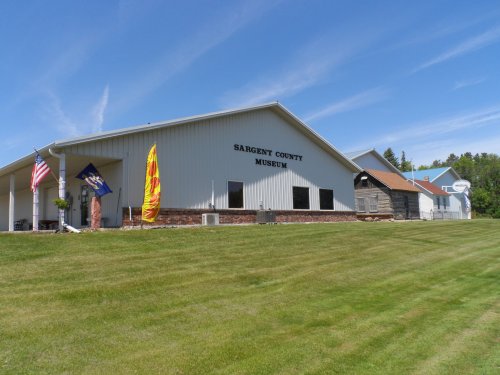 Exterior of the Sargent County Museum, featuring flags and a well-maintained lawn under a clear blue sky.