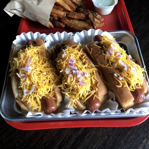 Three hot dogs topped with shredded cheese and diced onions, served with a side of fries and dipping sauce.