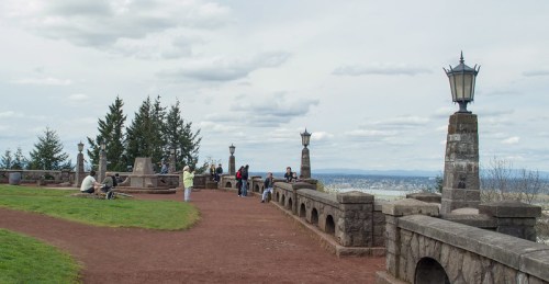 People enjoying a scenic overlook with stone walls and lanterns, surrounded by trees and cloudy skies.