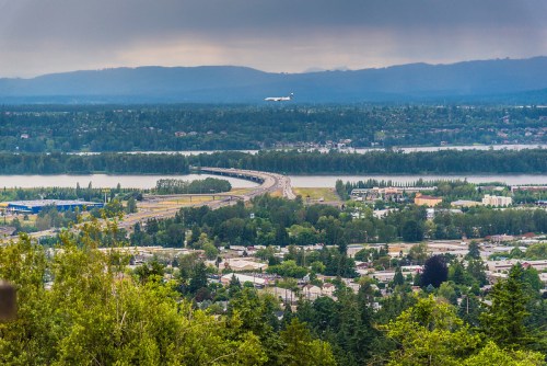 Aerial view of a highway, river, and cityscape with an airplane flying above in a cloudy sky.