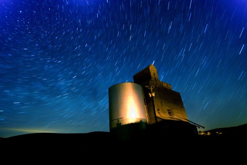 A grain silo under a starry night sky, with star trails creating a circular pattern around it.