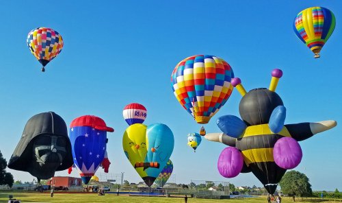 Colorful hot air balloons in various shapes, including a bee and a character, floating against a clear blue sky.
