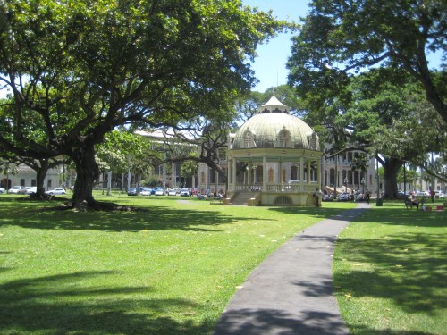 A gazebo surrounded by lush green grass and trees, with a pathway leading through a sunny park.