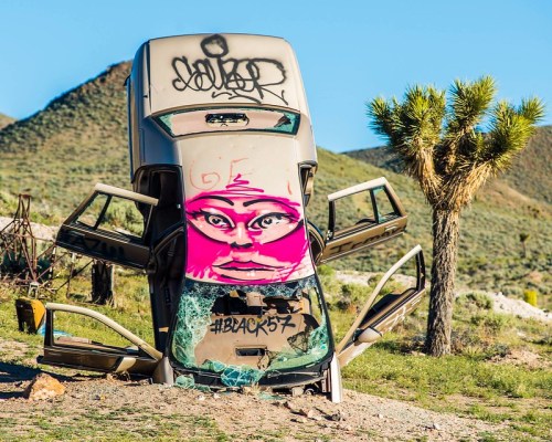 A graffiti-covered car is tilted on its side in a desert landscape, with a palm tree and mountains in the background.