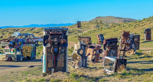A landscape of overturned, colorful cars in a grassy area, with mountains in the background under a clear blue sky.