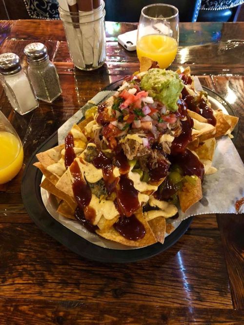 A large plate of nachos topped with pulled pork, guacamole, salsa, and barbecue sauce, served with drinks on the side.