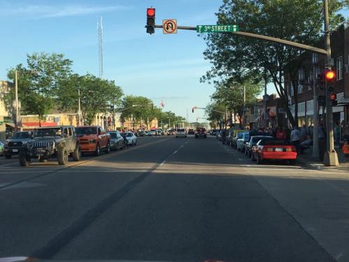 A busy street scene with parked cars, traffic lights, and people on the sidewalk under clear blue skies.
