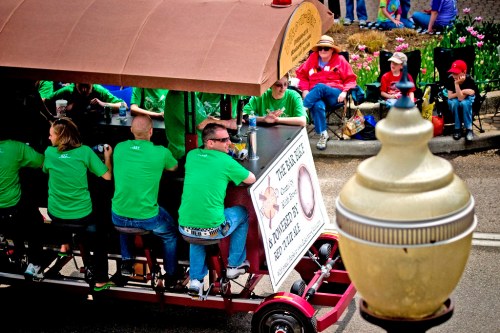 A group of people in green shirts enjoy a pedal-powered bar during a parade, with spectators in the background.