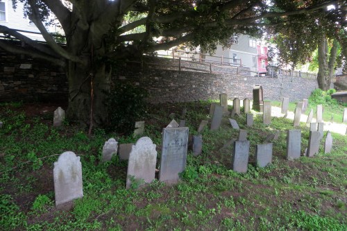 A cluster of weathered gravestones under a large tree, surrounded by grass and a stone wall in the background.