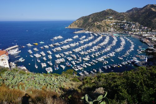 Aerial view of a marina filled with boats, surrounded by hills and clear blue water on a sunny day.