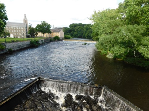 A river flows past a dam, with lush greenery and buildings visible along the banks under a clear sky.