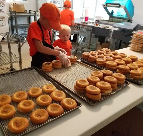 Two children in orange shirts are preparing donuts on trays in a kitchen.