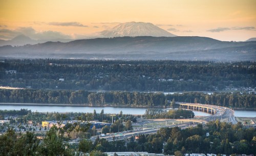 A scenic view of a river, bridge, and mountains under a sunset sky, with a snow-capped peak in the background.