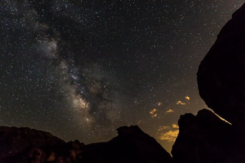 A starry night sky with the Milky Way visible, framed by dark rock silhouettes and soft clouds.