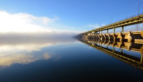 A serene view of a bridge reflecting in calm water, with fog and blue sky in the background.