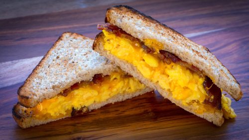 A toasted sandwich filled with scrambled eggs, melted cheese, and crispy bacon, cut in half on a wooden surface.