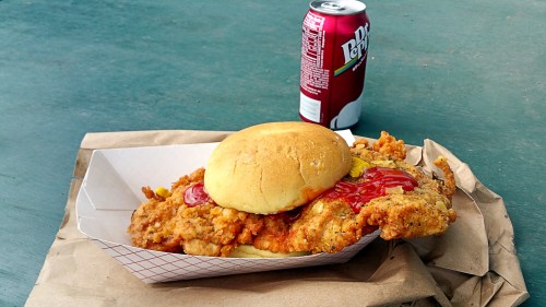 A fried chicken sandwich with ketchup and pickles on a paper tray, next to a can of soda on a green table.