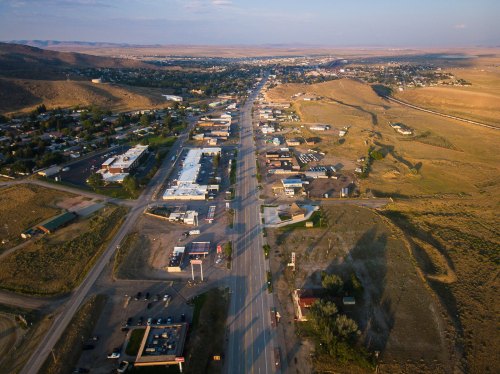 Aerial view of a small town with a main road, buildings, and surrounding hills under a clear sky.